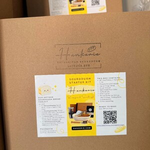 May include: A square brown cardboard box featuring a centered black logo for Hankerie Reimagined Sourdough. A white rectangular label with yellow accents lists the Sourdough Starter Kit contents, including a whisk, scraper, and proofing basket.