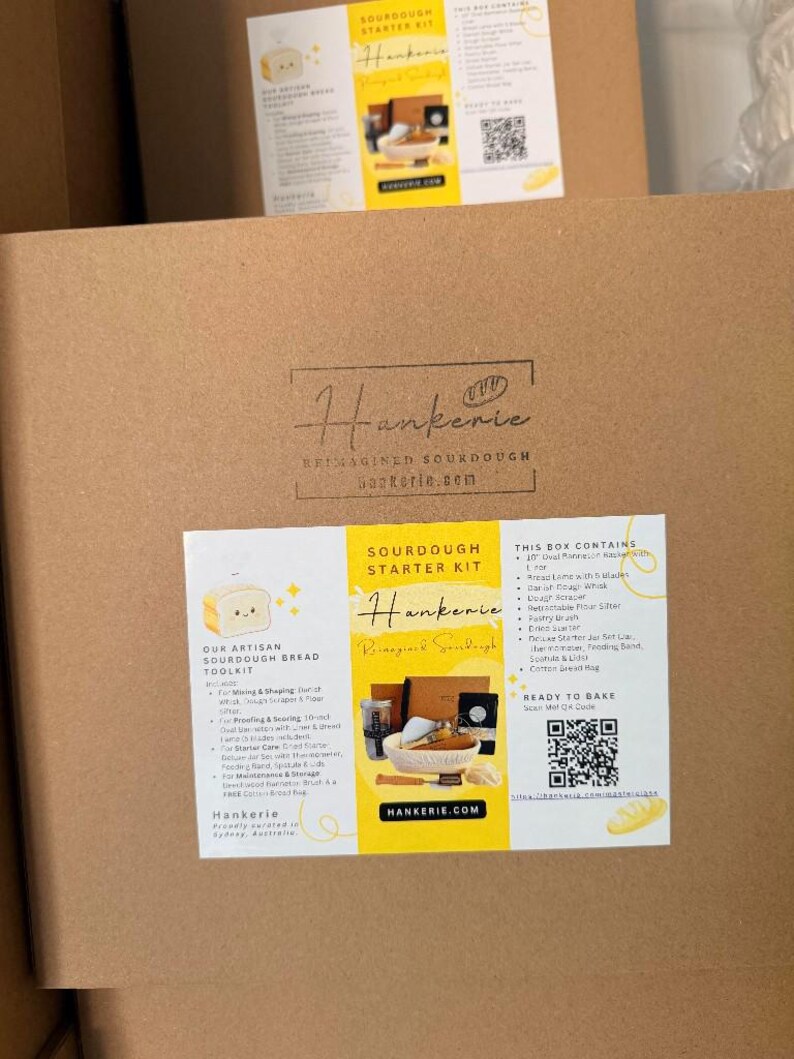 May include: A square brown cardboard box featuring a centered black logo for Hankerie Reimagined Sourdough. A white rectangular label with yellow accents lists the Sourdough Starter Kit contents, including a whisk, scraper, and proofing basket.
