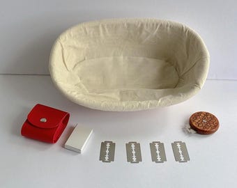 May include: A bread proofing basket with a cream-colored liner, a red leather pouch, a small silver box, four razor blades, and a wooden lid with a carved floral design. The items are arranged on a white surface.