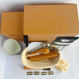 May include: A sourdough bread baking kit featuring a beige oval proofing basket, a wooden handled lame with four razor blades, a metal flour duster, a wooden brush, a round tin, two brown cardboard boxes, and a small beige cloth bag.