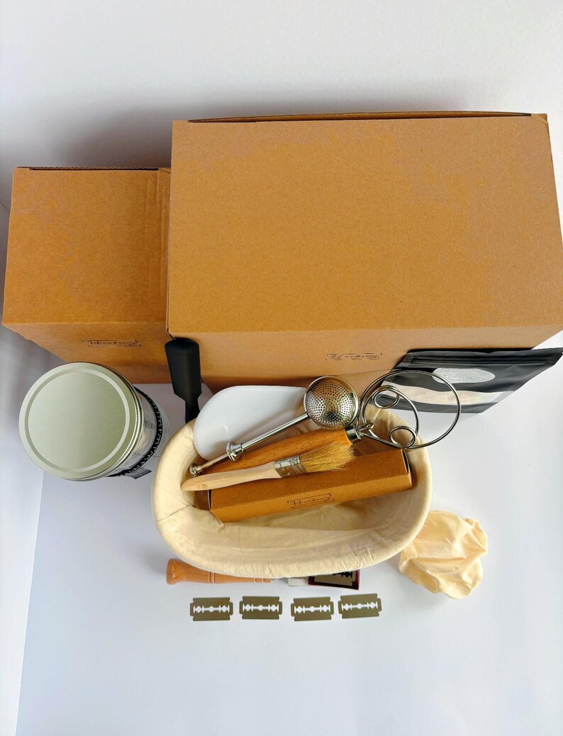 May include: A sourdough bread baking kit featuring a beige oval proofing basket, a wooden handled lame with four razor blades, a metal flour duster, a wooden brush, a round tin, two brown cardboard boxes, and a small beige cloth bag.