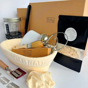 May include: A sourdough starter kit featuring a beige proofing basket, a glass jar with measurement markings, a black pouch labeled Hankerie, a metal whisk, a wooden brush, a dough scraper, a lame with a blade, and extra razor blades.