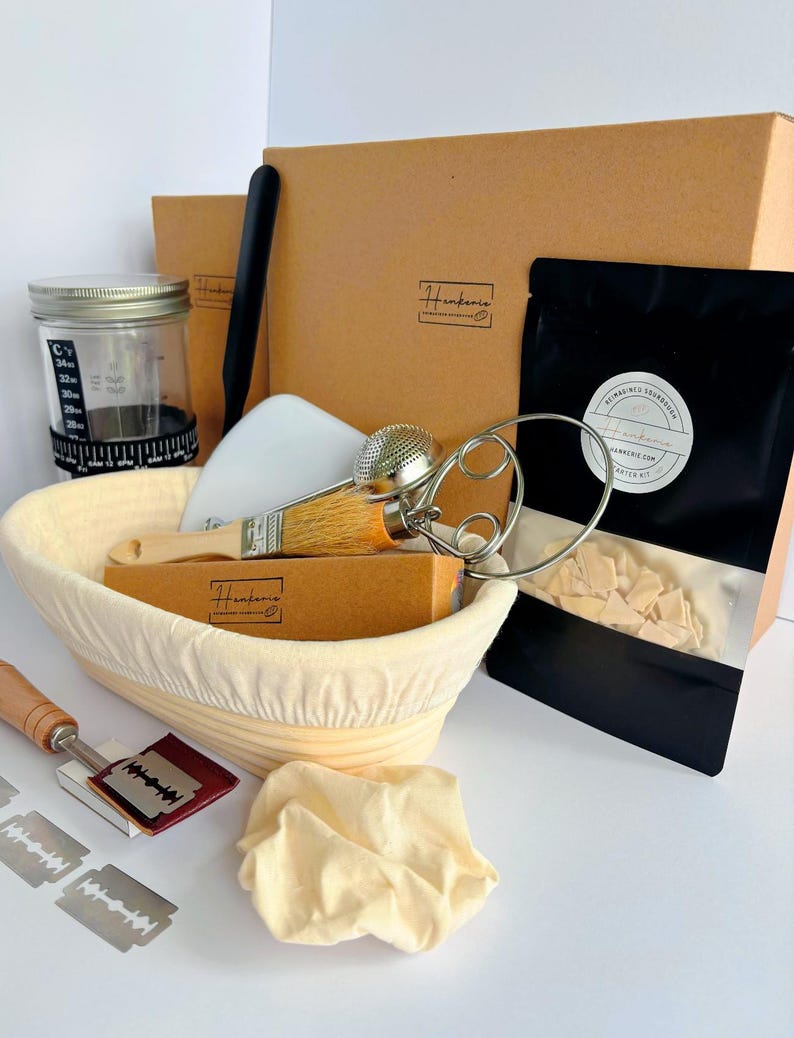 May include: A sourdough starter kit featuring a beige proofing basket, a glass jar with measurement markings, a black pouch labeled Hankerie, a metal whisk, a wooden brush, a dough scraper, a lame with a blade, and extra razor blades.
