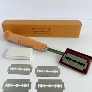 May include: A bread lame with a light wood handle and a metal blade attached to a dark red leather cover. The set includes four double-edged stainless steel razor blades, a small white box, and a rectangular brown cardboard box labeled Henkenis.