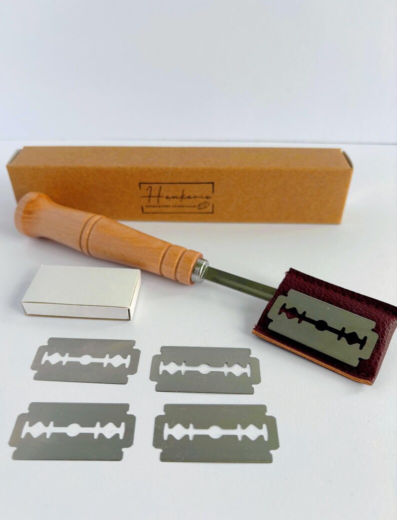 May include: A bread lame with a light wood handle and a metal blade attached to a dark red leather cover. The set includes four double-edged stainless steel razor blades, a small white box, and a rectangular brown cardboard box labeled Henkenis.