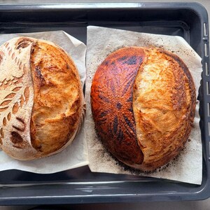 May include: Two round sourdough loaves of bread resting on parchment paper inside a dark gray rectangular baking tray. The left loaf features intricate leaf and heart scoring patterns, while the right loaf has a dark, toasted floral design.