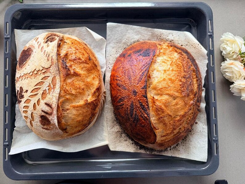 May include: Two round sourdough loaves of bread resting on parchment paper inside a dark gray rectangular baking tray. The left loaf features intricate leaf and heart scoring patterns, while the right loaf has a dark, toasted floral design.