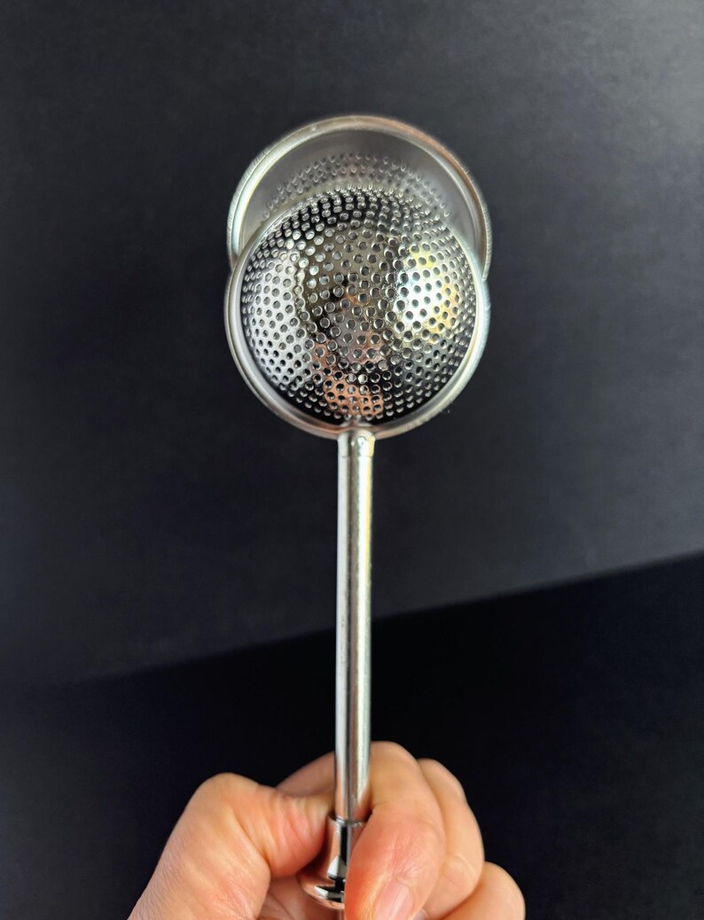 May include: A silver-colored metal tea infuser featuring a spherical, perforated mesh head attached to a straight, slender handle. The circular mesh component is designed to hold loose tea leaves, with a polished metallic finish throughout.