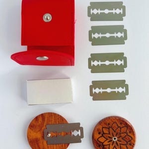 May include: A red leather case with a silver snap closure, a small white rectangular box, four silver double-edge razor blades, and two circular wooden discs. One disc features a laser-engraved floral pattern, while the other holds a blade.