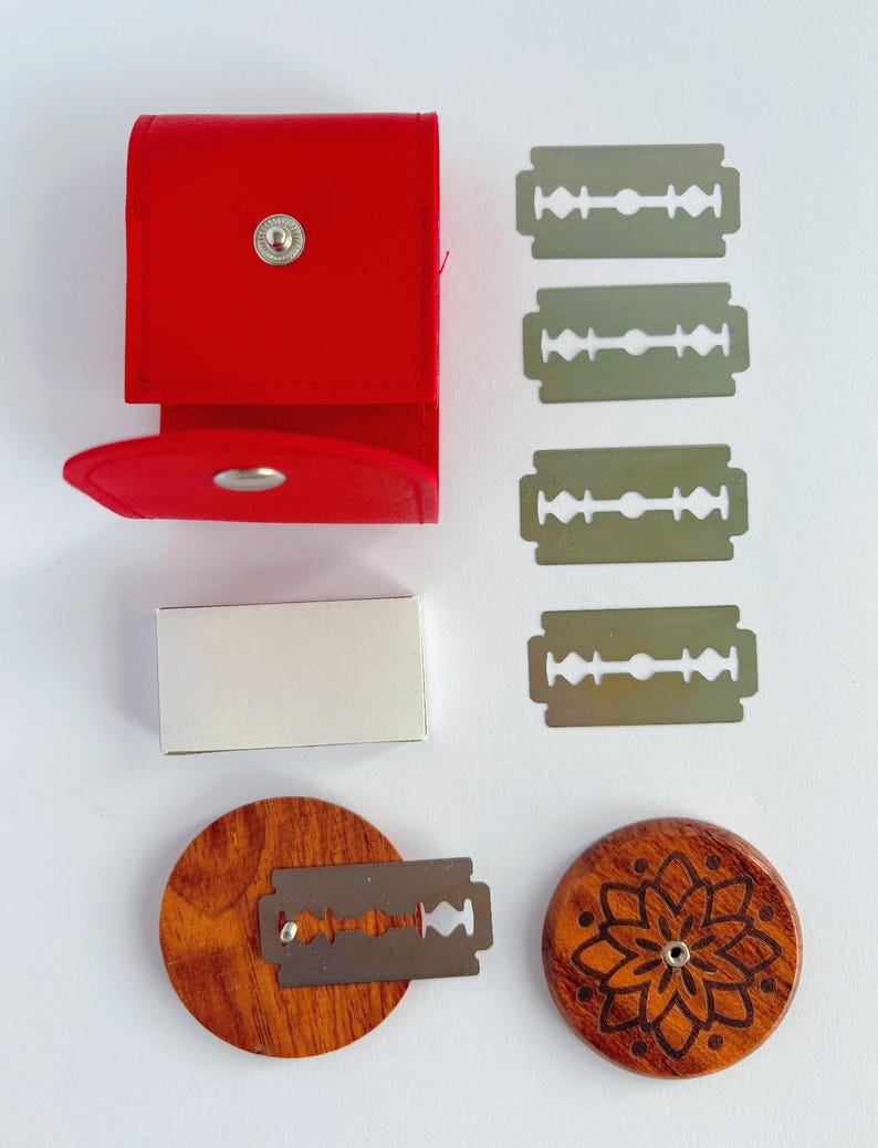 May include: A red leather case with a silver snap closure, a small white rectangular box, four silver double-edge razor blades, and two circular wooden discs. One disc features a laser-engraved floral pattern, while the other holds a blade.