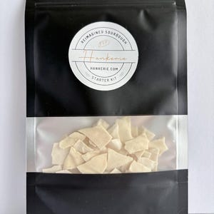 May include: A black resealable pouch with a clear window containing off-white, dried sourdough flakes. A circular white label features the text REIMAGINED SOURDOUGH, Hankerie, HANKERIE.COM, and STARTER KIT, along with a small bread icon.