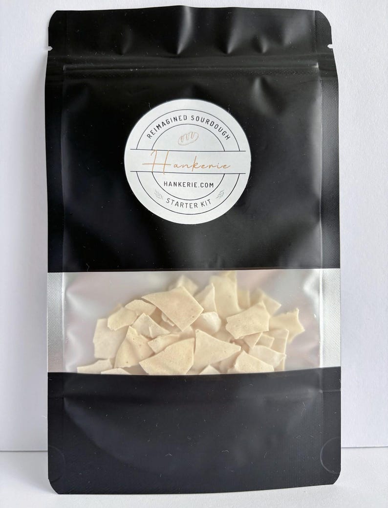 May include: A black resealable pouch with a clear window containing off-white, dried sourdough flakes. A circular white label features the text REIMAGINED SOURDOUGH, Hankerie, HANKERIE.COM, and STARTER KIT, along with a small bread icon.