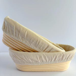 May include: Two oval-shaped proofing baskets made of light-colored natural cane, stacked one inside the other. Each basket is lined with a fitted, off-white fabric cover that drapes over the top rim, showing the ribbed texture of the wood.