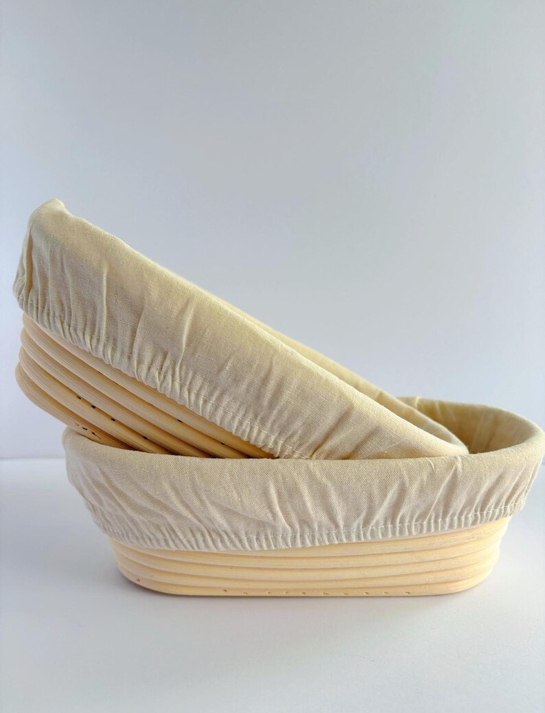 May include: Two oval-shaped proofing baskets made of light-colored natural cane, stacked one inside the other. Each basket is lined with a fitted, off-white fabric cover that drapes over the top rim, showing the ribbed texture of the wood.