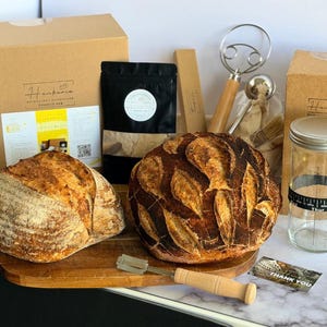 May include: A sourdough bread making kit featuring two round loaves of crusty bread, a wooden-handled scoring tool, a glass jar with a black date-tracking band, a black pouch, a wire dough whisk, and cardboard boxes with Hankerie branding.