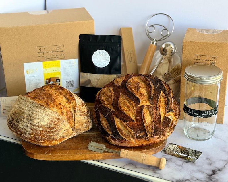 May include: A sourdough bread making kit featuring two round loaves of crusty bread, a wooden-handled scoring tool, a glass jar with a black date-tracking band, a black pouch, a wire dough whisk, and cardboard boxes with Hankerie branding.