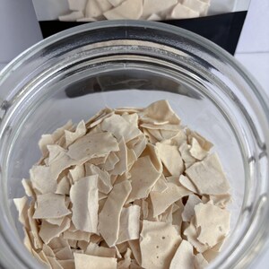 May include: A clear glass jar filled with small, irregular, off-white brittle shards. A matching plastic pouch containing more of the same light-colored, thin, jagged pieces is positioned directly behind the open glass container.
