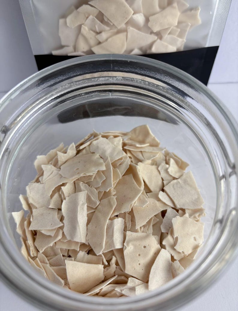 May include: A clear glass jar filled with small, irregular, off-white brittle shards. A matching plastic pouch containing more of the same light-colored, thin, jagged pieces is positioned directly behind the open glass container.