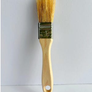 May include: A paint brush with light-colored natural bristles, a silver-toned metal ferrule stamped with the text 1 1/2 inch, and a smooth, light-colored wooden handle featuring a circular hole at the bottom for hanging storage.