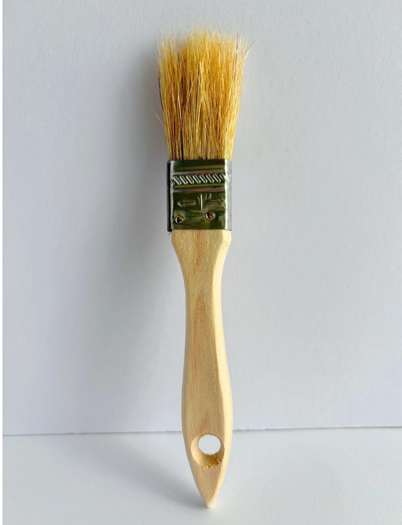 May include: A paint brush with light-colored natural bristles, a silver-toned metal ferrule stamped with the text 1 1/2 inch, and a smooth, light-colored wooden handle featuring a circular hole at the bottom for hanging storage.