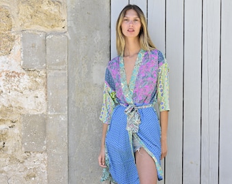 May include: A woman wearing a blue and green patterned kimono with a pink floral design. The kimono is tied at the waist with a matching sash. She is standing in front of a white wooden door.