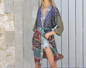May include: A woman wearing a long, multi-colored patterned kimono with a green sash. She is standing in front of a white wooden door with a stone wall behind her.