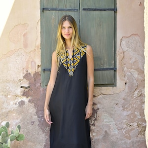 May include: A black sleeveless maxi dress with a geometric pattern in yellow and white on the neckline. The dress is worn by a woman standing in front of a wall with a green wooden door.