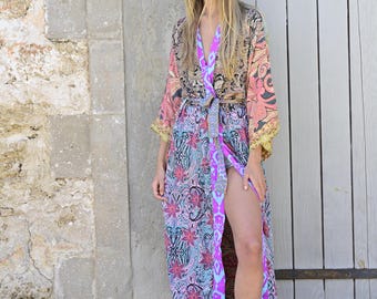 May include: A long, colorful floral print robe with a pink sash and a long, flowing silhouette. The robe is worn by a person standing in front of a wooden door.