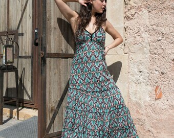 May include: A woman wearing a long, flowing maxi dress with a teal and black geometric pattern. The dress has a tiered skirt and a V-neckline. She is standing next to a wooden door.