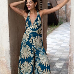 May include: A teal jumpsuit with a halter neckline and wide legs. The jumpsuit features a gold, stylized floral pattern. The model is standing in an outdoor setting, leaning against a wall.