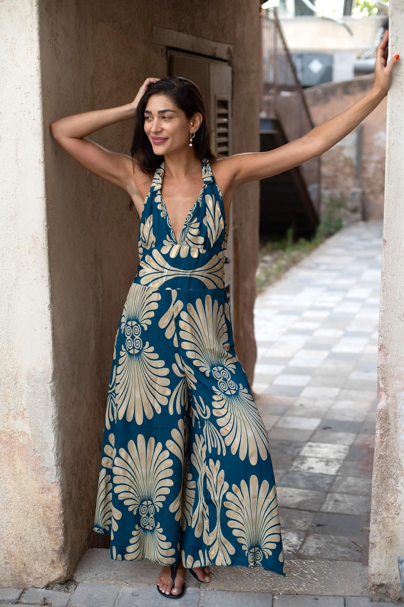 May include: A teal jumpsuit with a halter neckline and wide legs. The jumpsuit features a gold, stylized floral pattern. The model is standing in an outdoor setting, leaning against a wall.