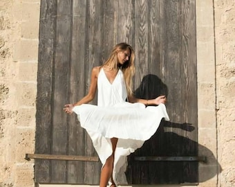 May include: A woman wearing a white, flowing dress with a V-neckline and spaghetti straps. The dress is knee-length and is being held up, revealing the legs. The background features a weathered wooden door and a stone wall.