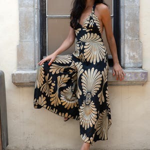 May include: Black halter-neck jumpsuit with a gold and cream-colored floral pattern. The wide-leg jumpsuit features a V-neckline and is worn with black sandals. The outfit is set against a neutral background.