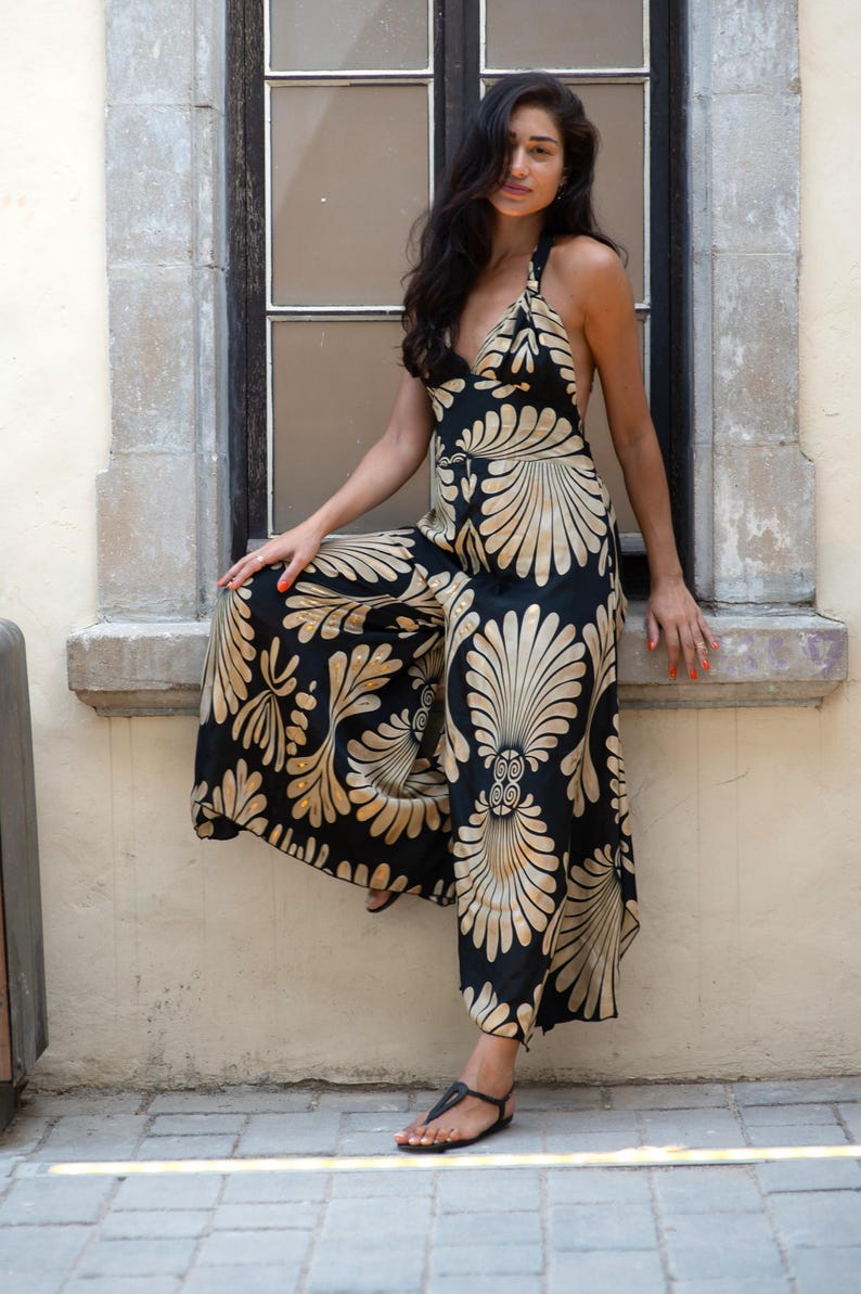 May include: Black halter-neck jumpsuit with a gold and cream-colored floral pattern. The wide-leg jumpsuit features a V-neckline and is worn with black sandals. The outfit is set against a neutral background.
