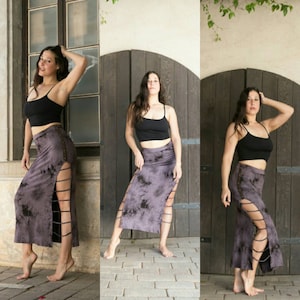May include: Three women wearing long, black and grey tie-dye skirts with side slits. The skirts have a unique design with multiple straps and buckles. The women are standing in front of a wooden door.