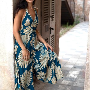 May include: A teal halter-neck jumpsuit with a cream-colored floral pattern. The wide-legged jumpsuit features a V-neckline and a flowing silhouette. The model is standing in an outdoor setting, leaning against a wall.