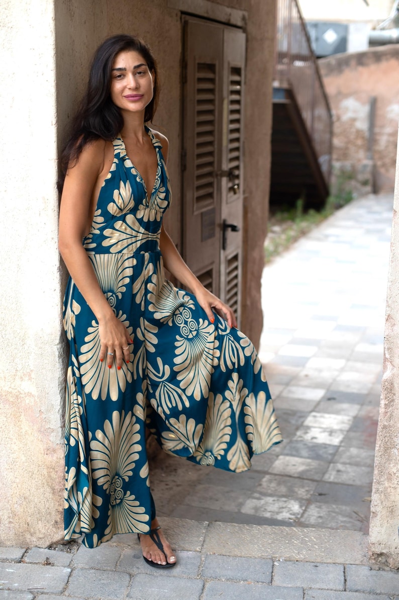 May include: A teal halter-neck jumpsuit with a cream-colored floral pattern. The wide-legged jumpsuit features a V-neckline and a flowing silhouette. The model is standing in an outdoor setting, leaning against a wall.