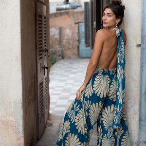 May include: A teal jumpsuit with a cream-colored floral pattern. The wide-leg pants and halter top are paired with a matching scarf tied at the back. The model is wearing black sandals.