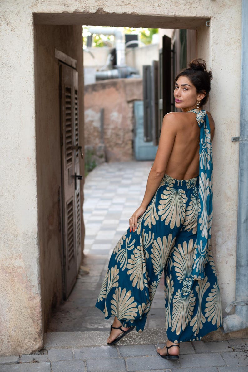 May include: A teal jumpsuit with a cream-colored floral pattern. The wide-leg pants and halter top are paired with a matching scarf tied at the back. The model is wearing black sandals.