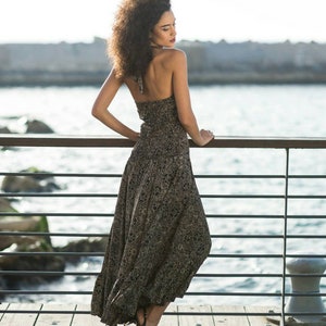 May include: A woman with curly hair wearing a black and brown patterned maxi dress and black heels stands on a wooden dock with a metal railing. The water is visible behind the railing.