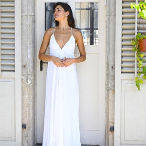 May include: A woman wearing a white maxi dress with a plunging neckline and thin straps. She is standing in front of a white door with shutters on either side.