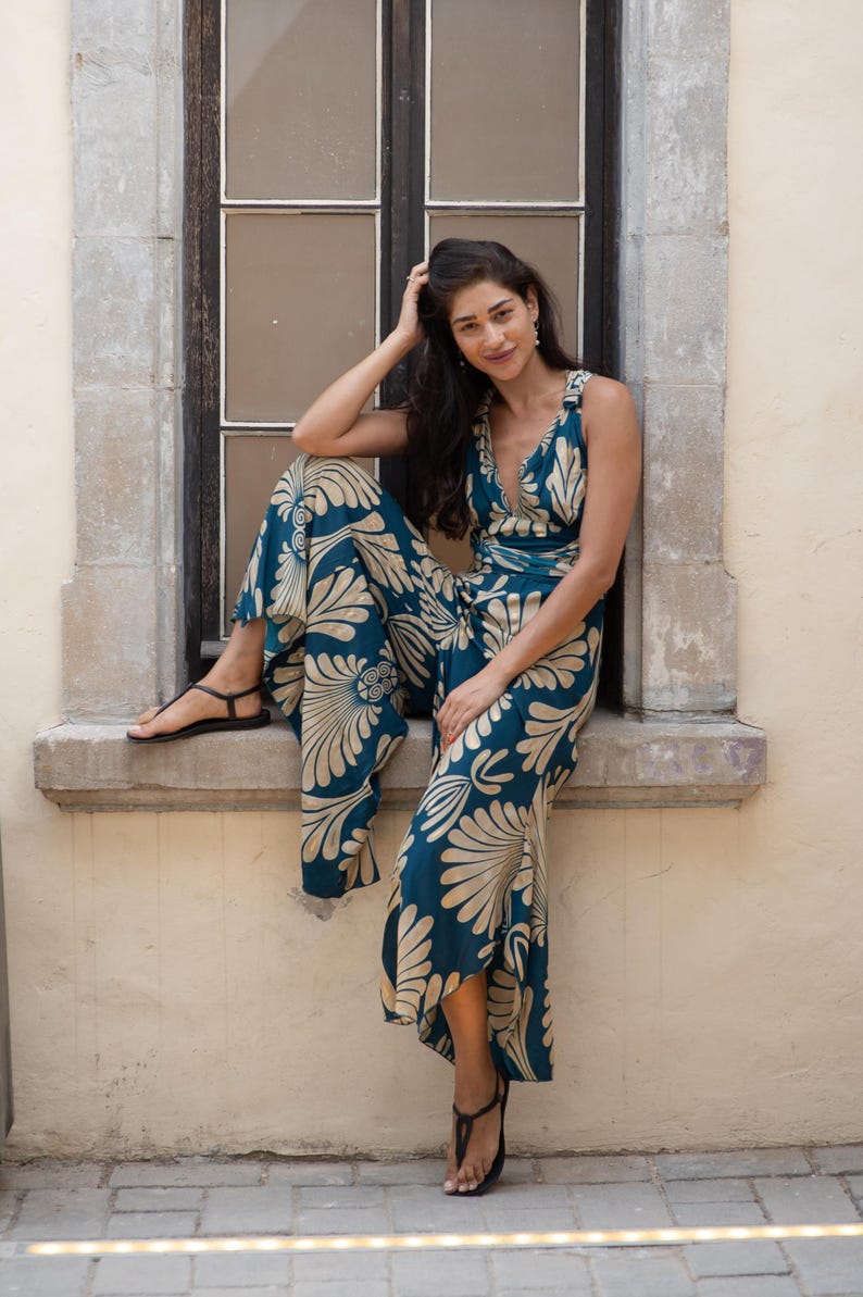May include: A sleeveless jumpsuit in teal with a cream-colored floral pattern. The jumpsuit features a V-neckline and wide legs. The model is wearing black sandals. The background is a window and a light-colored wall.