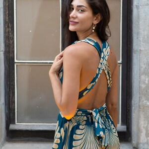 May include: A teal and cream-colored jumpsuit with a floral pattern. The backless design features crisscross straps and a bow at the waist. The model is standing in front of a window.