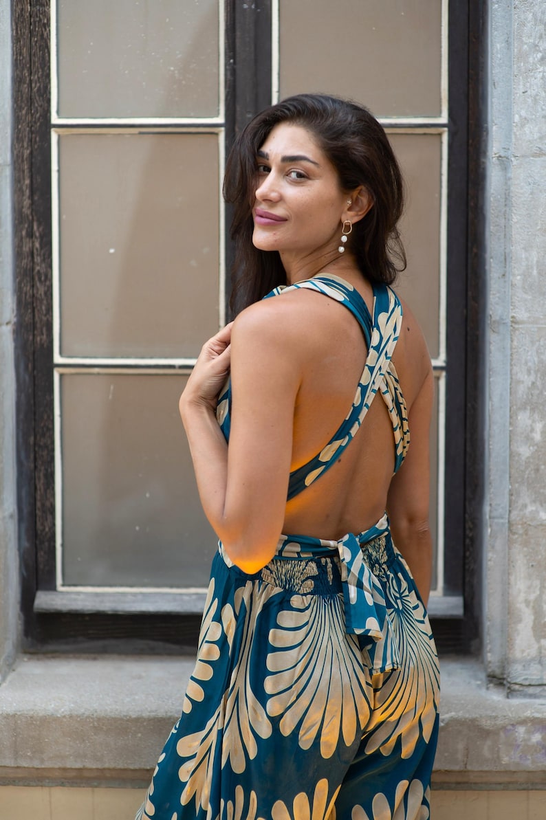 May include: A teal and cream-colored jumpsuit with a floral pattern. The backless design features crisscross straps and a bow at the waist. The model is standing in front of a window.