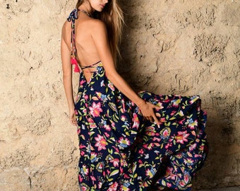 Op de afbeelding: Een vrouw draagt een blauwe maxi-jurk met een bloemenprint en roze kwastjes. De jurk heeft een halternek en een lage rug. Ze staat voor een stenen muur.