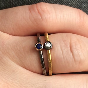May include: Two thin gold and black rings with small gemstones. The ring on the left has a blue gemstone and the ring on the right has a white gemstone.