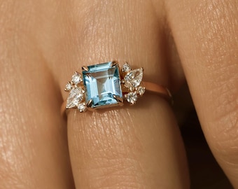 May include: A gold ring with a light blue square gemstone surrounded by smaller diamonds. The ring is on a finger.