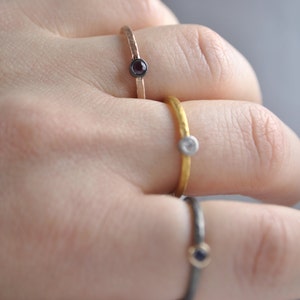 May include: Three simple gold rings with small gemstones. The rings are stacked on a finger. The top ring has a white gemstone, the middle ring has a black gemstone, and the bottom ring has a blue gemstone.