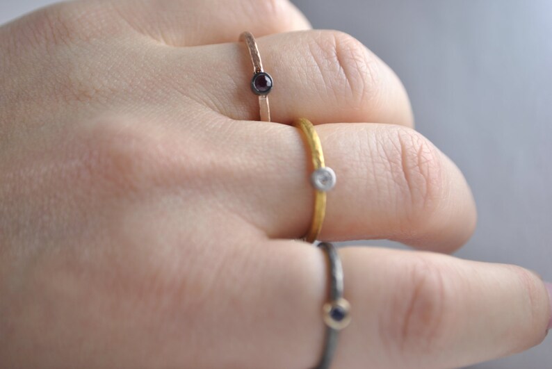 May include: Three simple gold rings with small gemstones. The rings are stacked on a finger. The top ring has a white gemstone, the middle ring has a black gemstone, and the bottom ring has a blue gemstone.