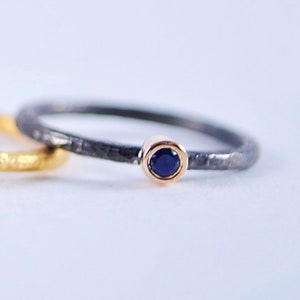 May include: A thin black ring with a hammered texture and a small blue gemstone set in a gold bezel.